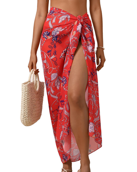 A woman models a red floral wrap sarong with a high side slit and knotted waist, holding a woven straw tote bag against a plain white background.